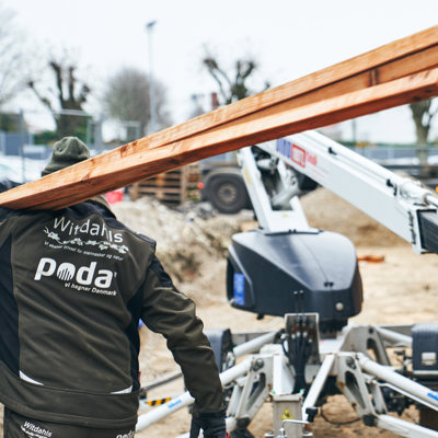 Monteur trägt lange Holzbretter auf einer Baustelle mit Hebebühne und Material.