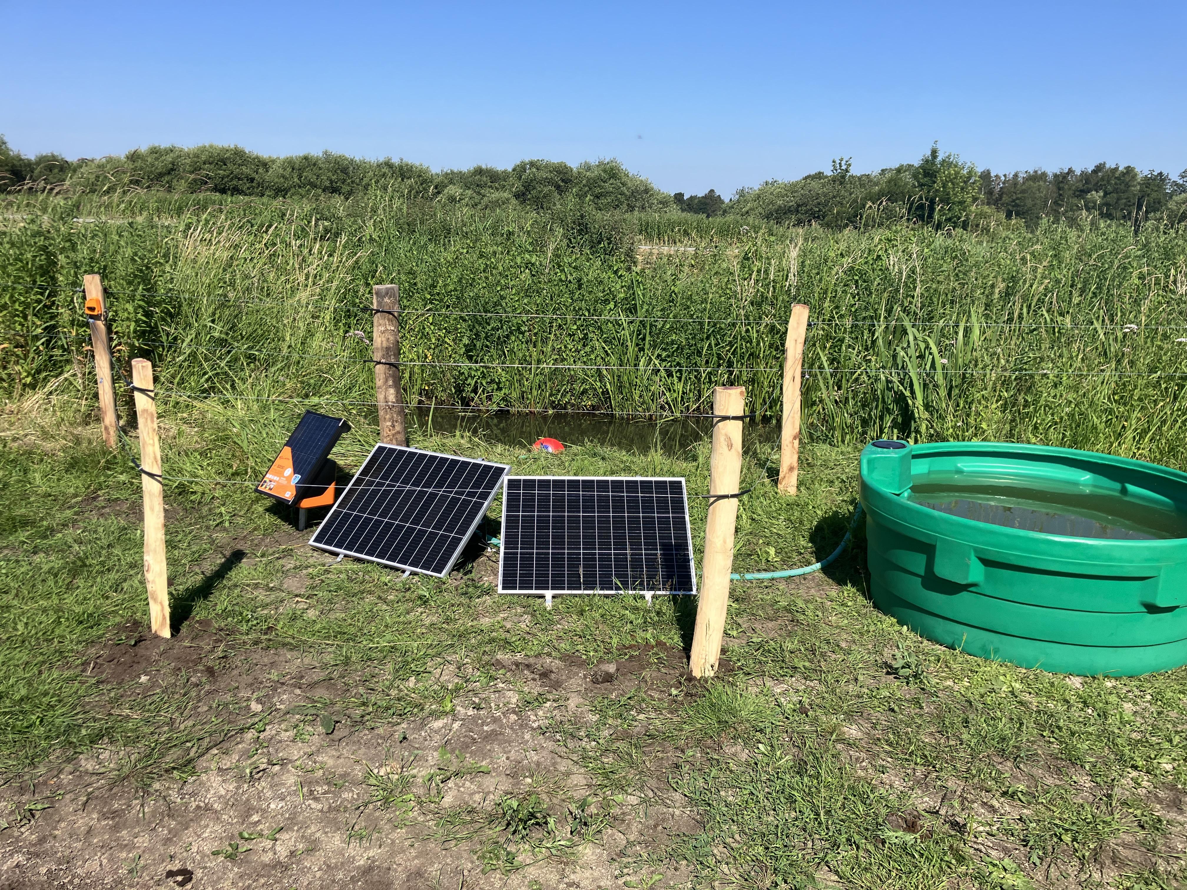Solarbetriebene Feldpumpe mit Solarpaneelen und Tränkschale an einem eingezäunten Bach.