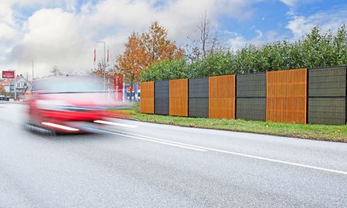 Noistop Essential Lärmschutzzaun mit einem schwarzen Rahmen und Metallpaneelen, auf denen einige Abschnitte mit optionalen Holzpaneelen versehen sind. Der Zaun ist von grüner Bepflanzung umgeben, und ein rotes Auto fährt schnell auf der Straße im Vordergrund vorbei.