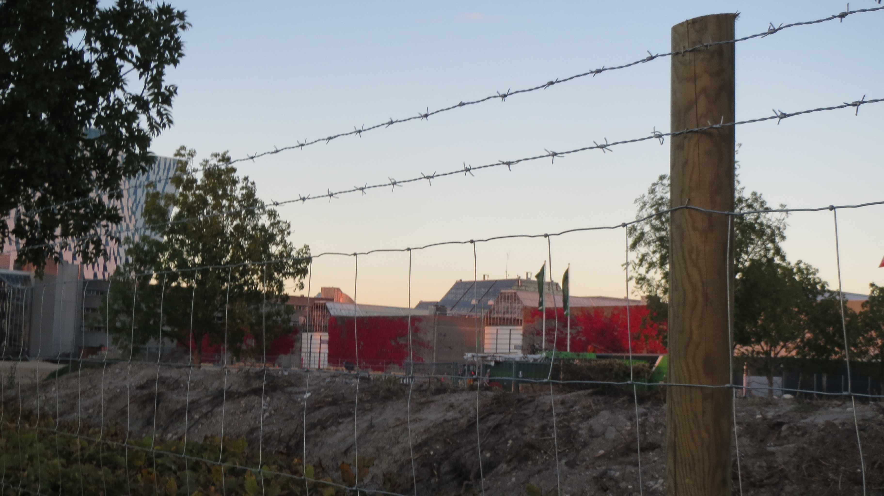 Im Hintergrund ist ein rotes Gebäude zu sehen, im Vordergrund ein Baustellenzaun