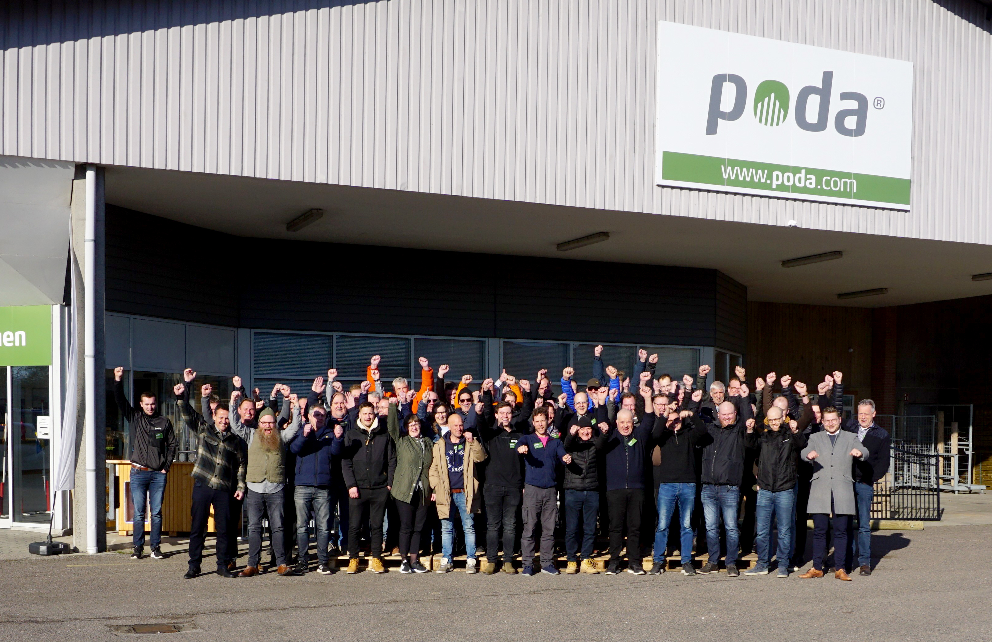 Gruppenfoto von Poda-Mitarbeitern vor dem Firmengebäude mit Poda-Logo und gemeinsam erhobenen Armen.