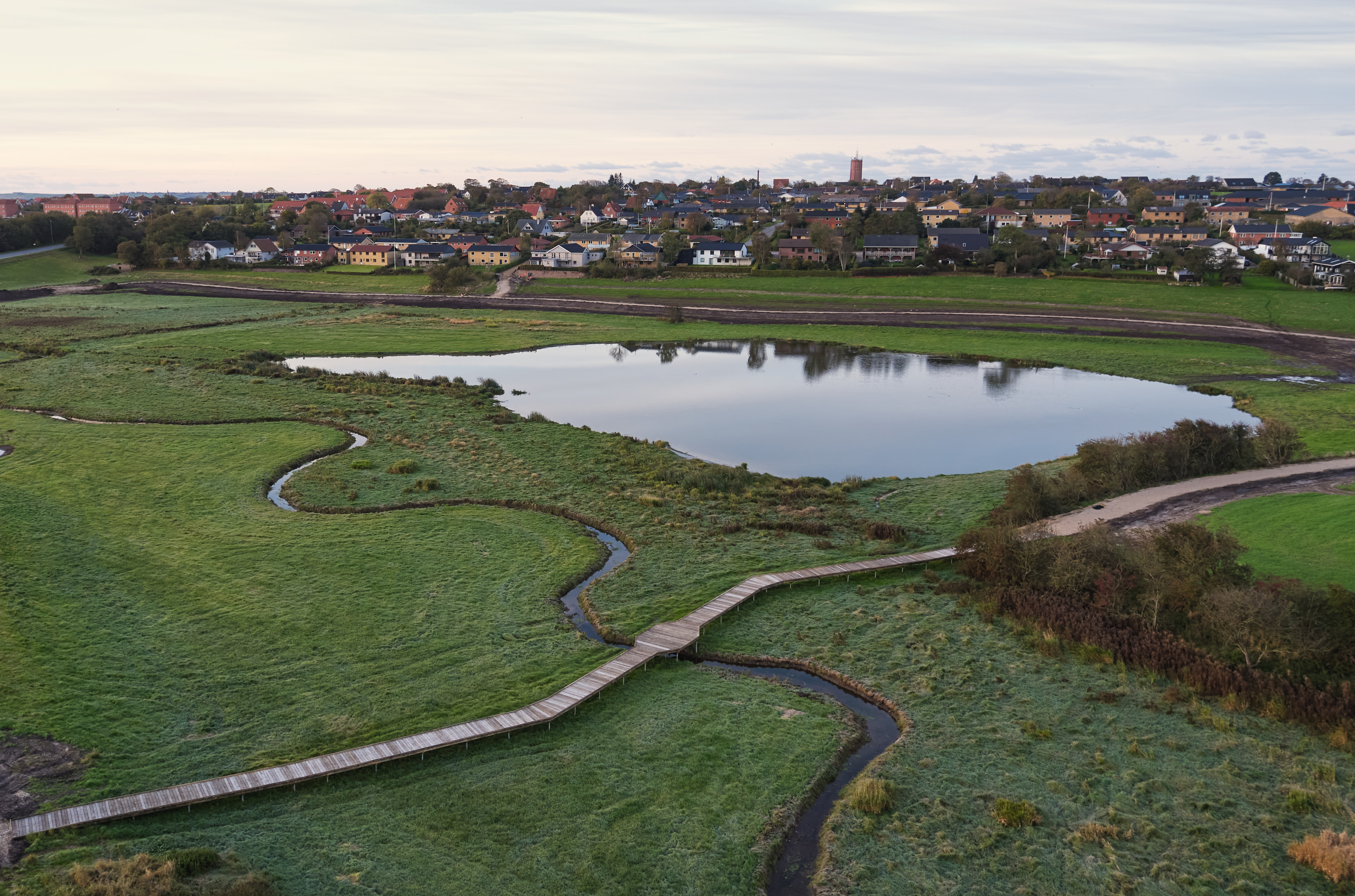 Naturschutzprojekt in einem sumpfigen Gebiet an einem See. Ein hölzerner Steg schlängelt sich durch die feuchte Landschaft und ermöglicht den Zugang, ohne die Natur zu stören. Im Hintergrund ist eine Stadt mit bunten Häusern und einer Kirche zu sehen.