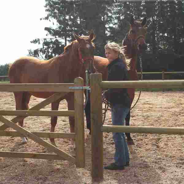 Eine Dame steht mit ihren beiden Pferden hinter einem Lattenzaun und einem Country Farmer- Tor