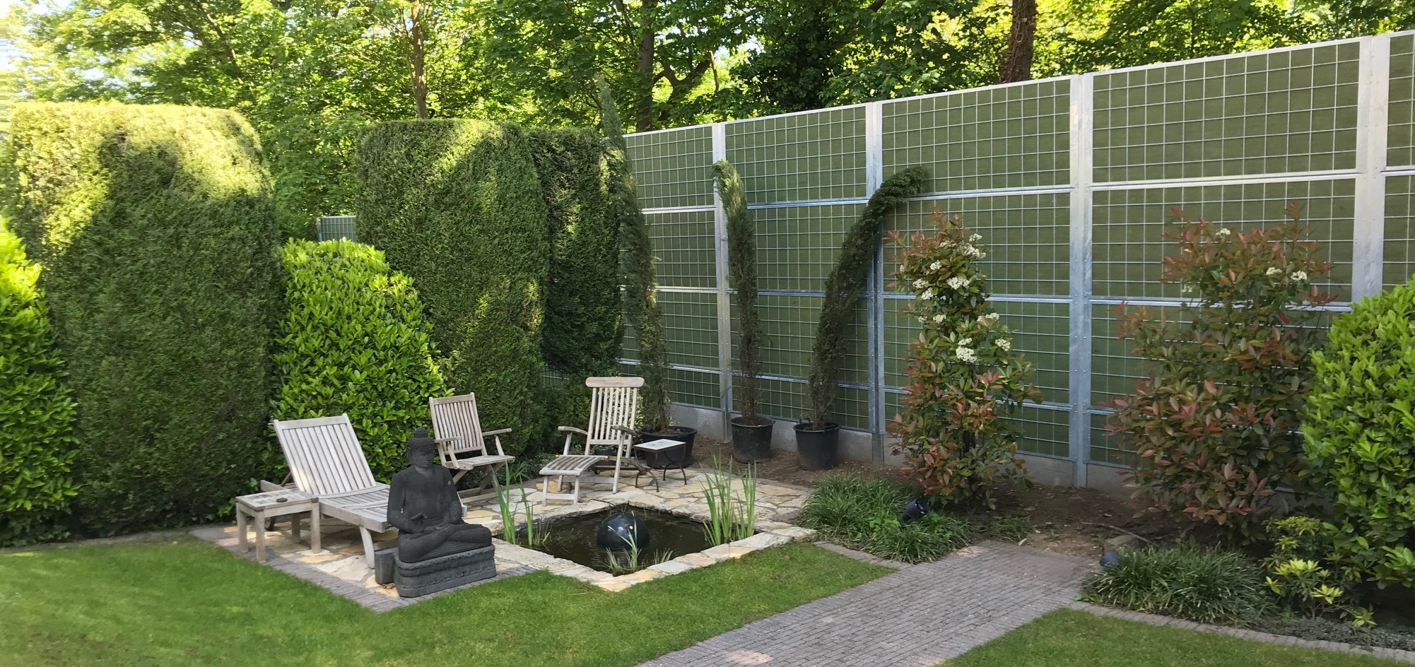Ein gemütlicher Garten mit Sträuchern und einem kleinen Fischteich, abgeschirmt vom Lärm der Umgebung mit einem Noistop® Steel-Lärmschutzzaun von Poda Zaun