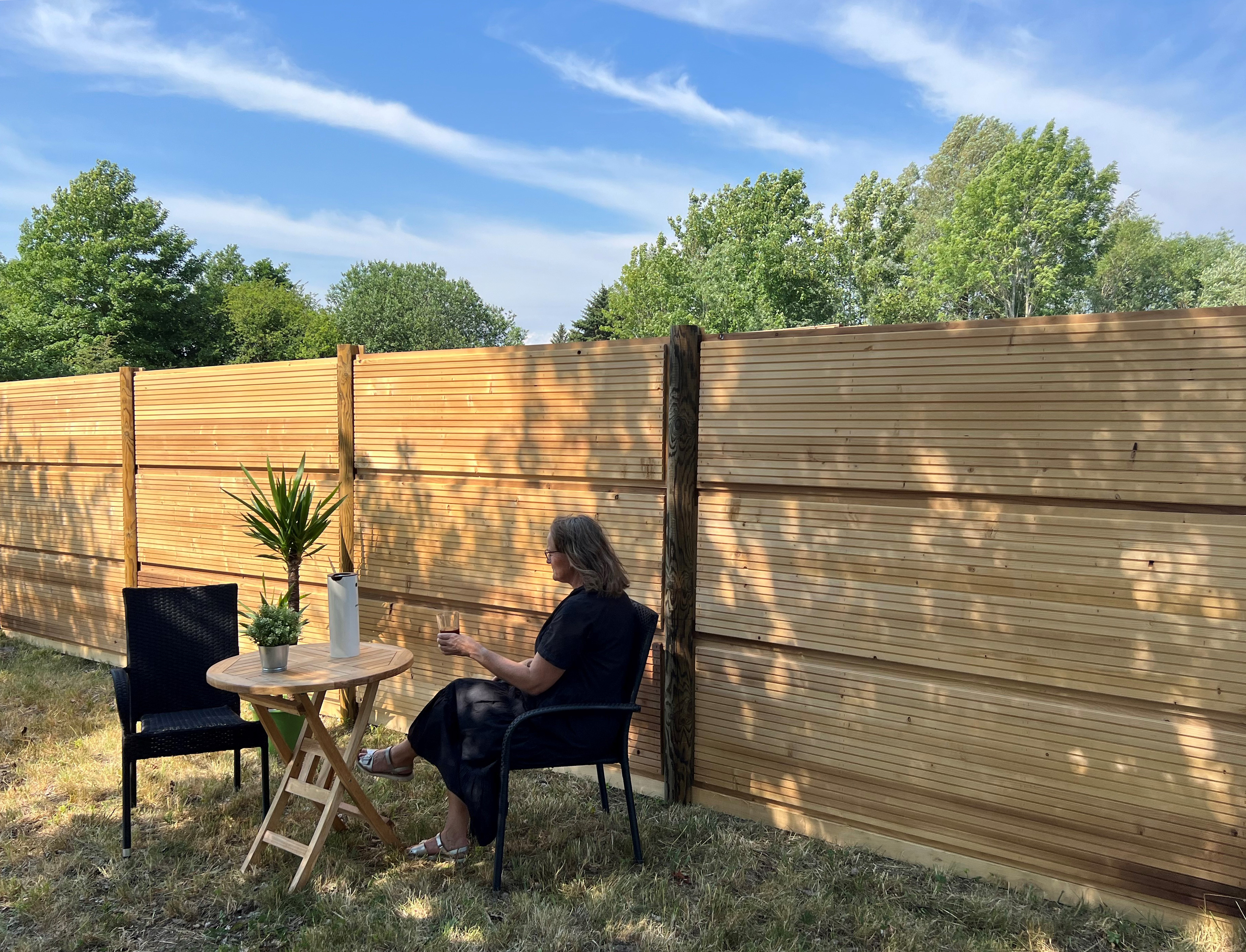 Eine Frau sitzt an einem Tisch mit einer Pflanze und einer Karaffe vor einer DeciFence-Holzschallschutzwand an einem sonnigen Tag.