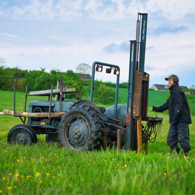 Monteur arbeitet mit einer Pfahlramme am Traktor beim Setzen von Zaunpfosten in einer grünen Feldlandschaft.