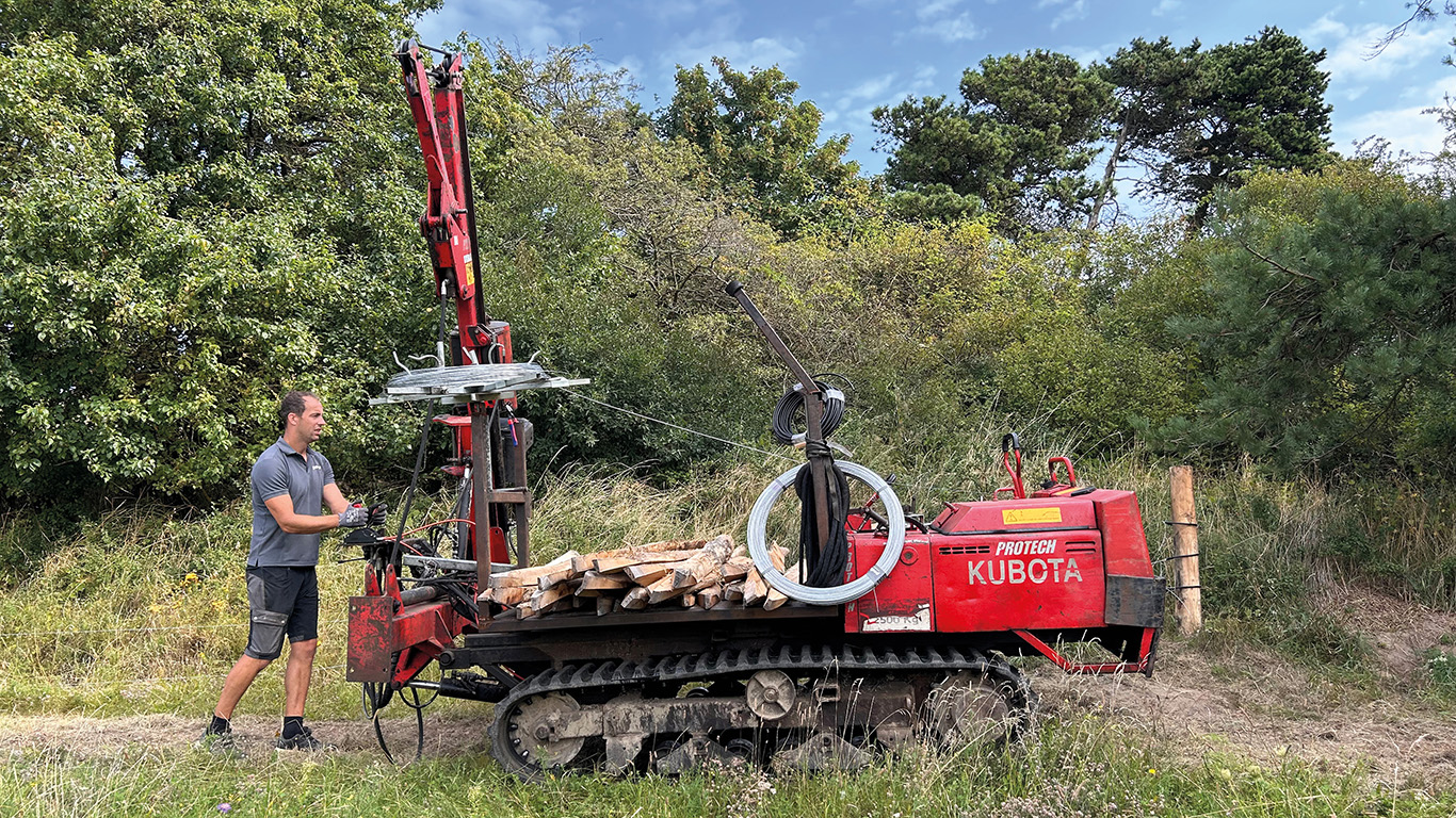Ein Monteur rollt Draht für einen Elektrozaun mit einer Spezialmaschine in einem grünen, waldreichen Gebiet ab.
