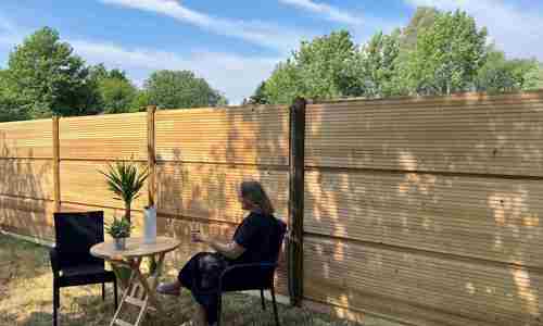 Eine Frau sitzt an einem Tisch mit einer Pflanze und einer Karaffe vor einer DeciFence-Holzschallschutzwand an einem sonnigen Tag.