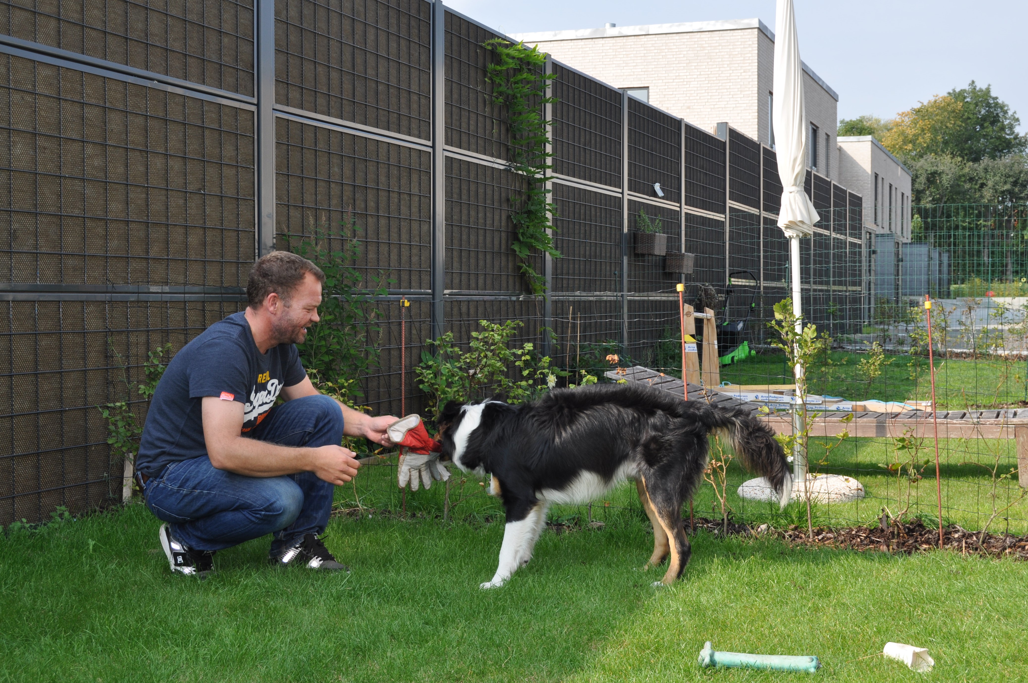 Ein Mann und sein Hund sitzen vor einem Noistop® Steel-Lärmschutzzaun von Poda Zaun, der in einem Garten aufgestellt ist, um den Lärm der Nachbarn abzuschirmen