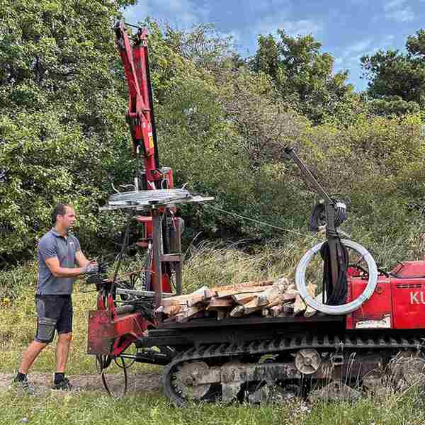 Ein Monteur rollt Draht für einen Elektrozaun mit einer Spezialmaschine in einem grünen, waldreichen Gebiet ab.