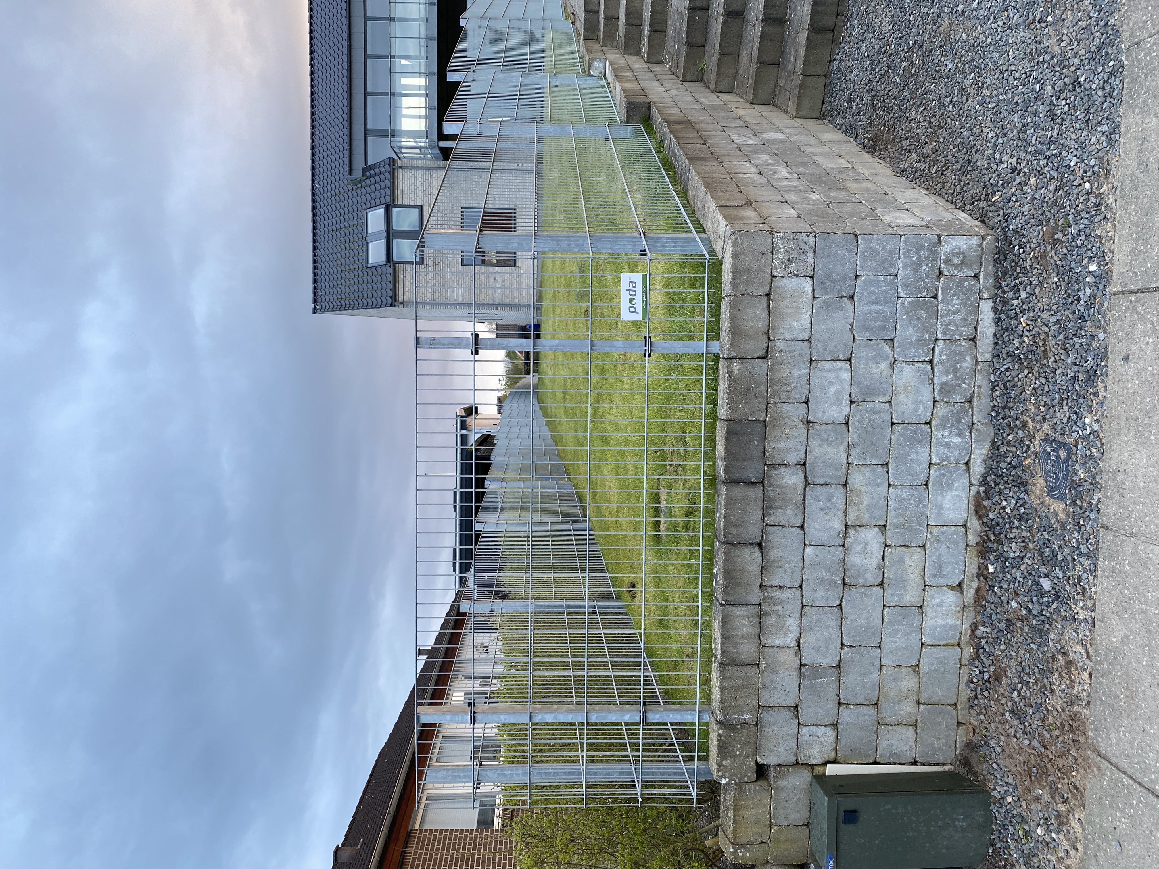 Nahaufnahme eines Doppelstabmattenzauns, der auf einer erhöhten Rasenfläche mit einer Stützmauer aus Betonblöcken montiert ist. Im Hintergrund ist ein modernes Haus mit großen Glasflächen zu sehen.