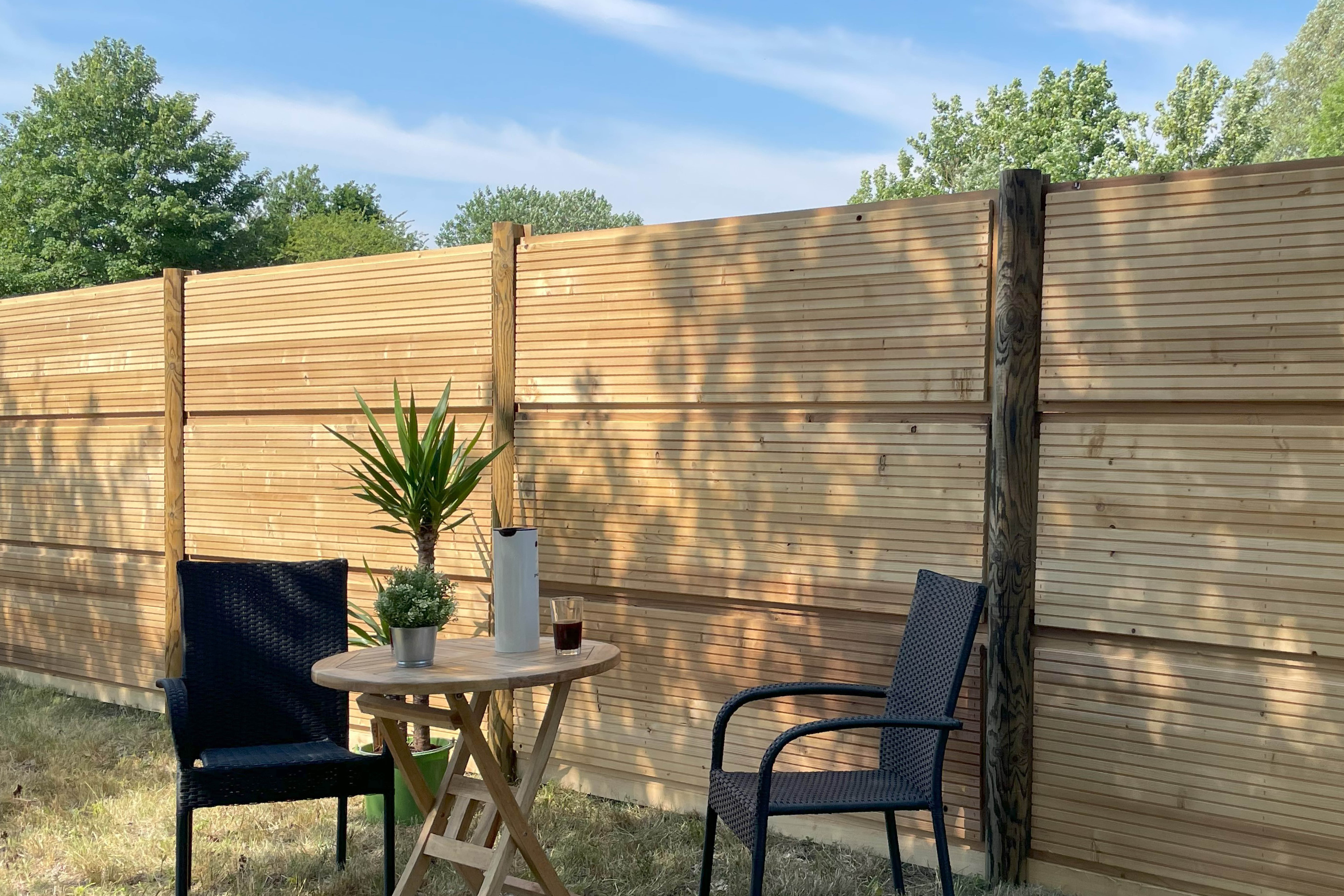 DeciFence Lärmschutzwand mit vertikalen Holzpfosten und horizontalen Paneelen. Vor dem Zaun steht eine Café-Garnitur mit zwei schwarzen Stühlen, einem kleinen runden Holztisch, einer Pflanze und einer Thermoskanne.