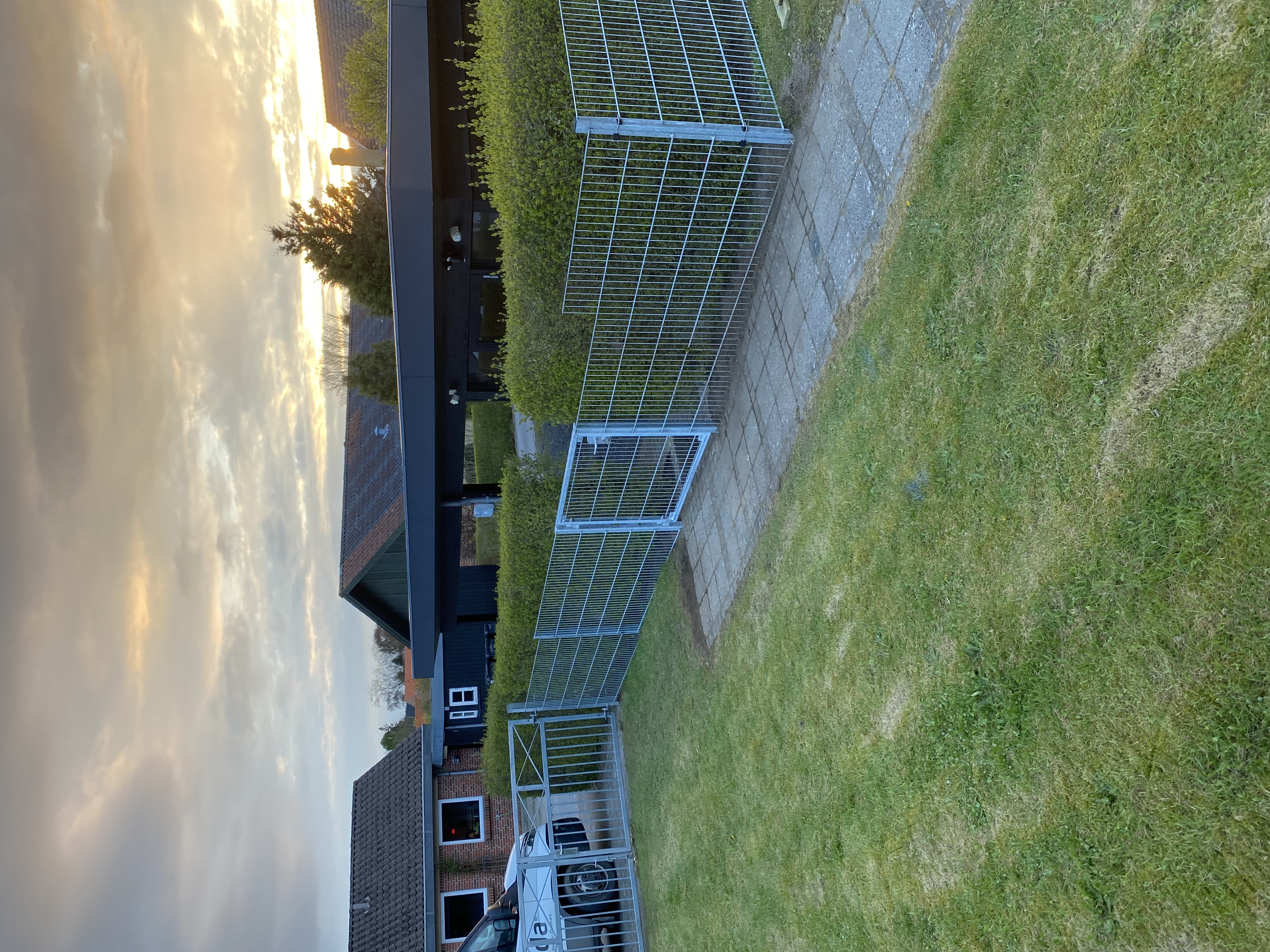 Doppelstabmattenzaun mit einem Tor entlang eines Weges, der eine Rasenfläche von einer Hecke trennt. Im Hintergrund sind ein Carport, mehrere Häuser und ein Sonnenuntergang zu sehen.